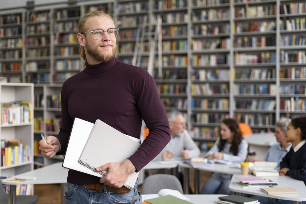 Stehender Mann in einer Bibliothek des hawvivaseo. Im Hintergrund sitzen vier Menschen an einem Tisch. Es sind Bücherregale zu sehen.